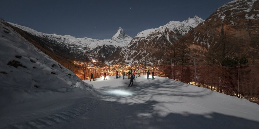 Piste in der Dunkelkeit mit Skifahrern, die auf das beleuchtete Zermatt hinunter blicken, im Hintergrund das Matterhorn | © Gabriel_Perren